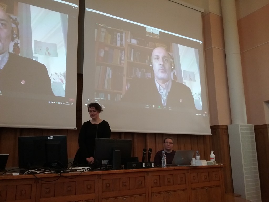 Me (Ellie) standing at a long desk looking at a screen. Next to me is Saana Svärd sitting behind a laptop at the same desk. Behind is a large screen showing Prof. Lorenzo Verderame during his statement about my dissertation.